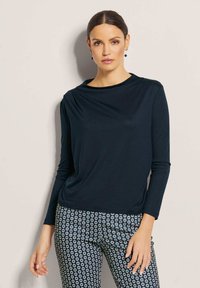 Marineblaues Langarmoberteil mit einem Bootsausschnitt, das über eine weiche Textur und einen lässigen Schnitt verfügt, kombiniert mit gemusterten blau-weißen Hosen.