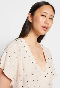 Blusa rosa clara com bolinhas pretas, detalhes de botões e um decote em V profundo. Mangas curtas e um tecido suave e texturizado.