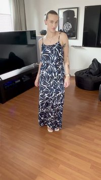 Une personne se tient dans le salon portant une robe longue sans manches à motif de feuilles noir et blanc et des sandales blanches. Télévision et pouf visibles en arrière-plan.