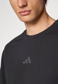 T-shirt à col rond noir en tissu lisse, avec un logo noir en relief discret sur le devant. Gros plan sur le col et la partie supérieure du torse.