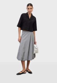 Mujer con camisa negra corta, falda midi gris texturizada, zapatos planos negros, sosteniendo un pequeño bolso plateado, de pie contra un fondo liso.