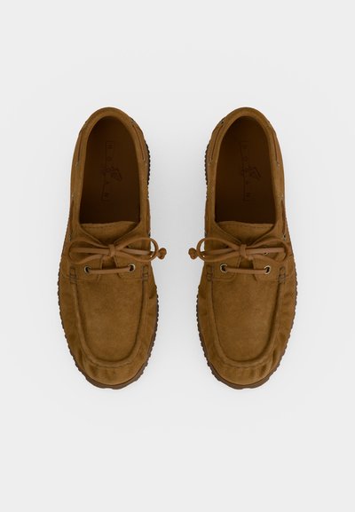Paire de mocassins en daim marron avec lacets en cuir noués en nœuds, vus de dessus sur fond blanc.