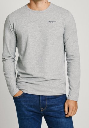 Mann trägt ein hellgraues Langarmshirt mit dem "Pepe Jeans London"-Logo auf der Brust und eine dunkelblaue Jeans, eine Hand in der Hosentasche.