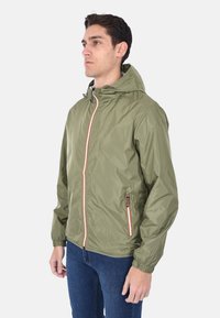 Giacca leggera verde oliva, con zip frontale e dettagli arancioni, cappuccio regolabile, polsini elasticizzati e tasca laterale con zip. Tessuto testurizzato.