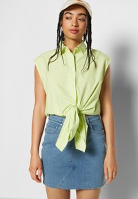 Camisola sem mangas verde-claro com detalhe frente atado, combinada com uma saia de ganga azul com bainha desfiada e costura diagonal.