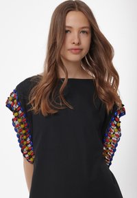 Zwarte jurk met kleurrijke kralenaccenten op de mouwen in blauw, rood, geel en groen, met een gladde, matte textuur.