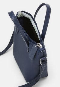 Borsa a tracolla sintetica blu navy con superficie testurizzata, chiusura con zip e tracolla regolabile. L'interno foderato presenta un'etichetta bianca con la scritta "LACOSTE."