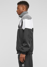 Black and white windbreaker jacket with a high collar, mesh detailing on the shoulders, elastic cuffs, and zippered pockets.