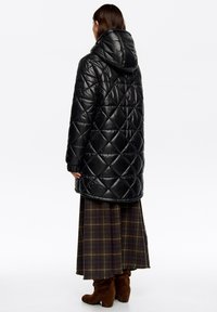 Czarna pikowana kurtka puchowa z kapturem, o błyszczącym wykończeniu i wzorze w romby, noszona na kraciastej spódnicy i brązowych zamszowych botkach.