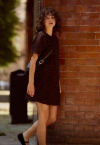 Leoprint-Kleid in Dunkelbraun und Schwarz, mit kurzen Ärmeln, lockere Passform. Getragen mit einer schwarzen Umhängetasche und schwarzen Ballerinas, vor einer Ziegelmauer posierend.
