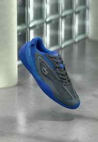 Αθλητικό παπούτσι Lacoste με γκρι επάνω μέρος με μπλε λεπτομέρειες, υφή δίχτυ, στρογγυλή μύτη και εύκαμπτη λαστιχένια σόλα. Μοντέλο LP003.