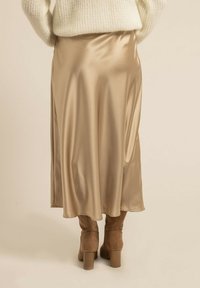 Jupe midi en satin beige, présentant une texture lisse et un drapé fluide. Portée avec des bottes beiges jusqu'aux genoux.