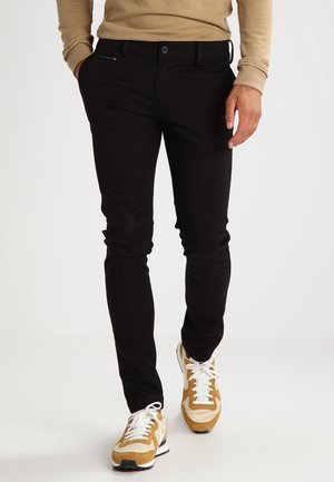 Uomo che indossa pantaloni neri aderenti, felpa beige e sneakers beige e bianche, che cammina con una mano in tasca.