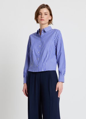 Femme portant une chemise à rayures verticales bleu et blanc à manches longues et un pantalon large foncé sur fond uni.