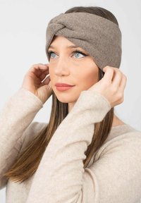 Bandeau en tricot marron avec un détail torsadé à l'avant. Le modèle ajuste le bandeau, portant un pull beige clair. Texture lisse, design décontracté.
