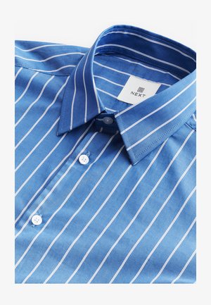 Zils kokvilnas krekls ar baltām svītrām, pogājamu apkakli un etiķeti ar uzrakstu "NEXT". Uzsver baltas pogas un gludu tekstūru.