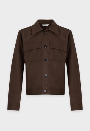 Jeune homme aux cheveux foncés coiffés en arrière, portant une veste marron boutonnée, un pantalon assorti, une chemise crème et des mocassins noirs, debout sur fond blanc.