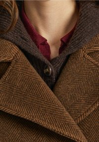 Cappotto marrone a spina di pesce con grandi revers, chiusura a pulsante e un colletto intrecciato marrone scuro sovrapposto a una camicia bordeaux.