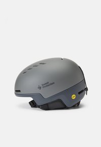 Casco gris con tecnología MIPS, acabado mate elegante, orificios de ventilación y correa de barbilla ajustable. Presenta un logo negro y un acento amarillo.
