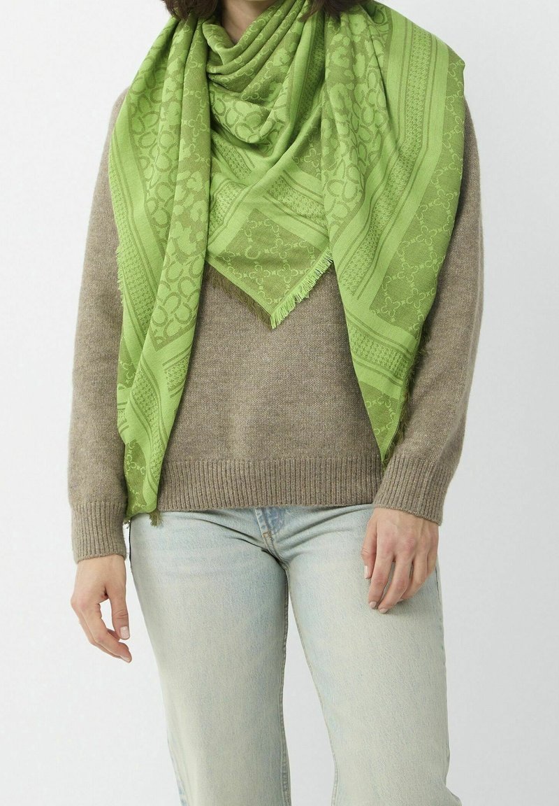 Personne portant un jean bleu clair, un pull en tricot beige et une grande écharpe verte à motifs drapée autour du cou et des épaules.
