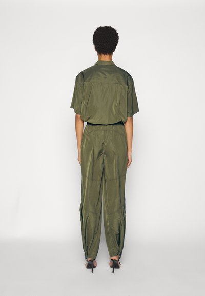 Olijfgroene twee-delige set bestaande uit een shirt met korte mouwen en getailleerde broek met ritsen bij de enkels en voorzakken. Soepele textuur.