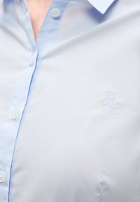 Svetlomodrá košeľa na zapínanie s hladkou látkou, ktorá má jemne vyšívané logo "MOP" a biele gombíky pozdĺž predného pásu.