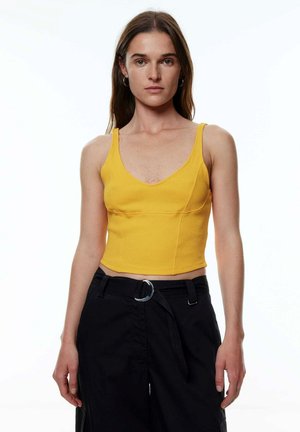 Geel geribbelde crop top met een diepe V-hals, voorzien van dunne bandjes. Gepaard met zwarte broek met een zilveren ring als ceintuur lus.