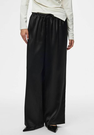 Femme portant un pantalon large en satin noir avec une ceinture élastique et des chaussures noires pointues, associé à un haut blanc à manches longues.