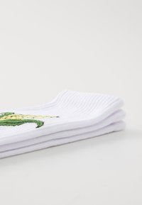 Chaussettes blanches en coton avec une texture côtelée, ornées d'un motif brodé d'une créature verte et d'accents jaunes sur le devant.