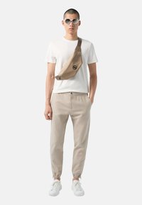 Sac bandoulière beige avec fermeture éclair et logo, t-shirt blanc, joggers beiges avec poignets élastiques, et baskets blanches. Des lunettes de soleil rondes complètent le look.