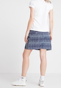 Niebieska sportowa spódnica w wzory z białymi paskami po bokach, zestawiona z gładką białą koszulką z krótkim rękawem i białymi butami sportowymi.