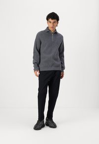Grau gestrickter Pullover mit einem Viertelreißverschluss-Kragen, strukturierte Oberfläche, kombiniert mit schwarzen Hosen und dunkel gemusterten Sneakers.