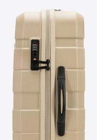 Valigia rigida beige con esterno testurizzato, chiusura con zip, lucchetto combinato integrato e manico nero. Forma rettangolare con scanalature.