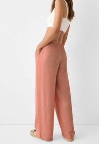Femme portant un pantalon corail ample taille haute avec poches, un crop top en crochet blanc et des chaussures beige à enfiler, se tenant de côté.