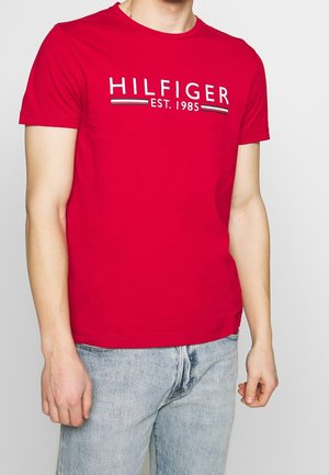 Man wearing a red short-sleeve t-shirt with "HILFIGER EST. 1985" text and light blue denim jeans, standing against a plain background.