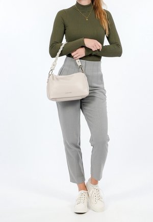 Kobieta ubrana w oliwkowozielony prążkowany golf, szare spodnie z wysokim stanem, białe trampki, trzymająca beżową torebkę na ramię z plecionym paskiem.