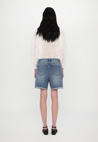 Blusa bianca leggera e trasparente indossata su pantaloncini di jeans blu con orlo sfilacciato. Il modello indossa scarpe flat nere con borchie.