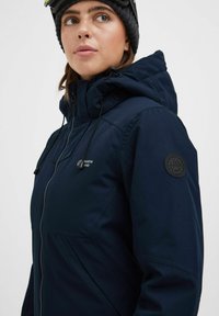 Tamsiai mėlynas lauko švarkas su užtrauktuku, reguliuojamu gaubtu, raišteliais ir logotipo etikete ant kairiojo rankovės. Lygi, vandeniui atspari medžiaga.