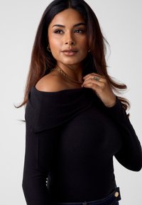 Zwart off-shoulder top met een strakke pasvorm, voorzien van een zachte textuur en lange mouwen. Gouden kettingaccent rond de halslijn.