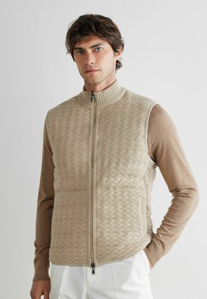 Mann trägt eine beige gesteppte Weste mit Reißverschluss über einem hellbraunen Langarmshirt und weißen Hosen, steht vor einem schlichten weißen Hintergrund.