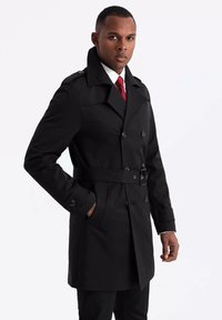 Czarny płaszcz trench z paskiem w talii, podwójnym zapięciem na guziki i dużymi kieszeniami bocznymi. Posiada szeroki kołnierz oraz fakturowaną tkaninę.