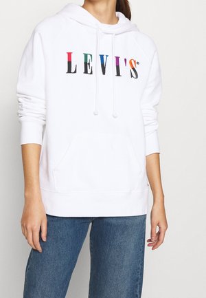 Osoba nosząca białą bluzę z kapturem z wielokolorowym logo "LEVI'S" na piersi i niebieskie dżinsy na jednolitym tle.