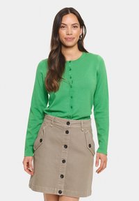 Camicia verde a maniche lunghe con bottoni e finitura testurizzata, abbinata a una gonna di jeans beige con tasche laterali e bottoni neri.