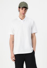 White cotton polo shirt with short sleeves and a collar, paired with black shorts. A green cap is worn on the head. Simple design, no patterns.