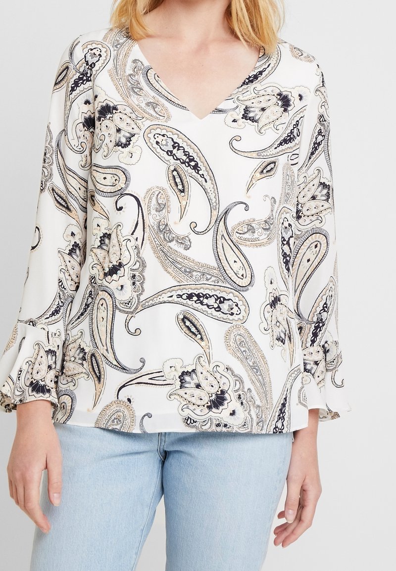 Femme portant un chemisier blanc à col en V avec des motifs paisley noirs et beiges et un jean bleu clair, montrée des épaules aux hanches.