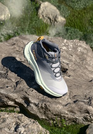 Scarpa da trekking grigio chiaro con tomaia in rete testurizzata, dettagli arancioni, colletto imbottito e suola flessibile, posizionata su una superficie rocciosa.