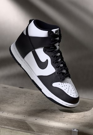 Baskets montantes Nike noir et blanc suspendues au-dessus d'une surface en béton avec des ombres douces en arrière-plan.