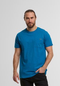 Blaues kurzärmliges T-Shirt aus weichem Stoff, mit einer Brusttasche und kleinen, mehrfarbigen Akzenten. Lässige Passform.