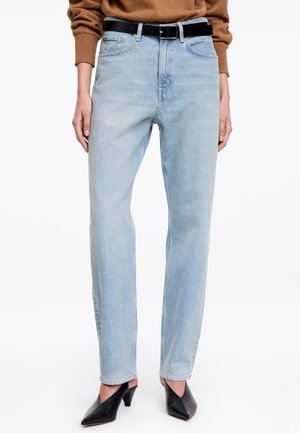 Personne portant un jean droit bleu clair avec une ceinture noire, un pull marron et des chaussures à talons pointues noires sur fond blanc.