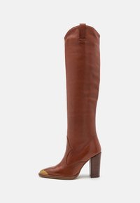 Bota alta de cuero marrón hasta la rodilla con punta afilada, detalle dorado en la punta y tacón de madera apilada. Presenta una textura suave y un diseño ajustado.